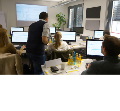 Schulungsraum mit mehreren Teilnehmenden an PCs, Trainer hilft einer Teilnehmerin