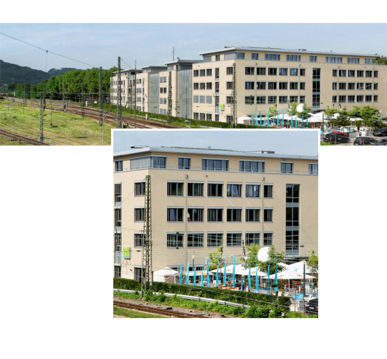Mehrstöckiges beiges Bürogebäude, Terrasse mit Sonnenschirmen, Bahngleise und Oberleitungen