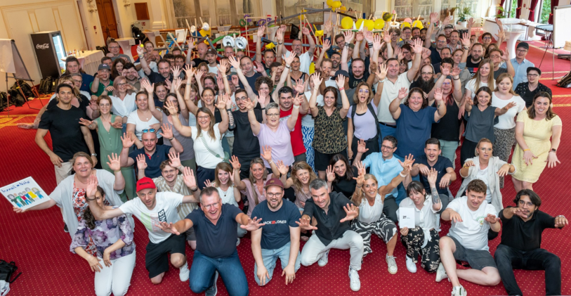 Gruppenfoto vieler Personen in einem Tagungsraum, alle winken zur Kamera
