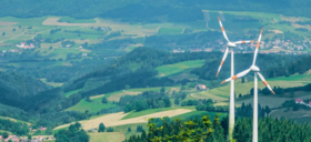 Zwei Windkraftanlagen vor hügeliger grüner Landschaft mit Feldern und Wäldern
