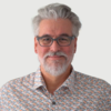 Smiling man with gray hair and beard, wearing glasses and a patterned shirt.