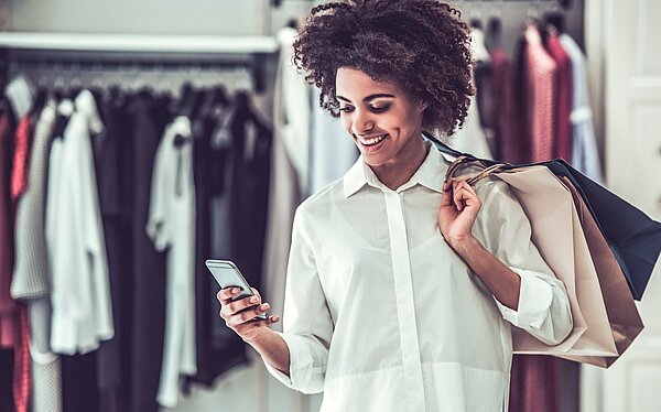 Eine lächelnde Frau in einem weißen Hemd steht in einem Bekleidungsgeschäft und hält Einkaufstaschen in der einen Hand und ein Smartphone in der anderen. Im Hintergrund sind Kleidungsstücke auf Kleiderbügeln zu sehen.