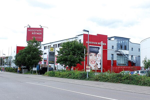 Außenansicht eines Modepark Röther-Gebäudes mit roter Fassade. Ein großes Werbeschild zeigt das Logo von Modepark Röther sowie eine Werbeanzeige mit einer lächelnden Frau. Im Vordergrund sind Bäume und parkende Autos zu sehen.