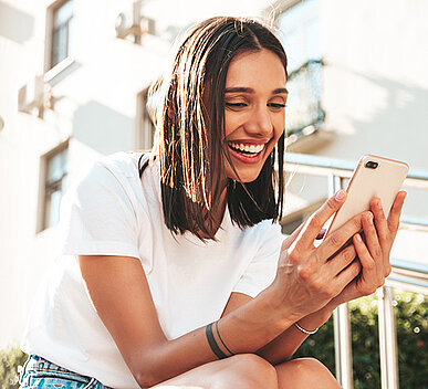 A woman smiling while looking at her smartphone, sitting outdoors on a sunny day.