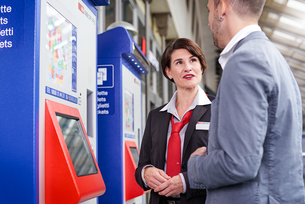 Eine Reiseberaterin der SBB steht an einem Fahrkartenautomaten und unterstützt einen Kunden beim Kauf von Tickets.