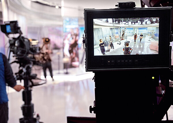 Eine Szene hinter den Kulissen einer Fernsehproduktion, bei der mehrere Kameras auf ein Set gerichtet sind. Auf einem Monitor ist die Live-Aufnahme des Sets zu sehen, auf dem mehrere Personen stehen, während im Hintergrund unscharfe Technik- und Studiomitglieder erkennbar sind.